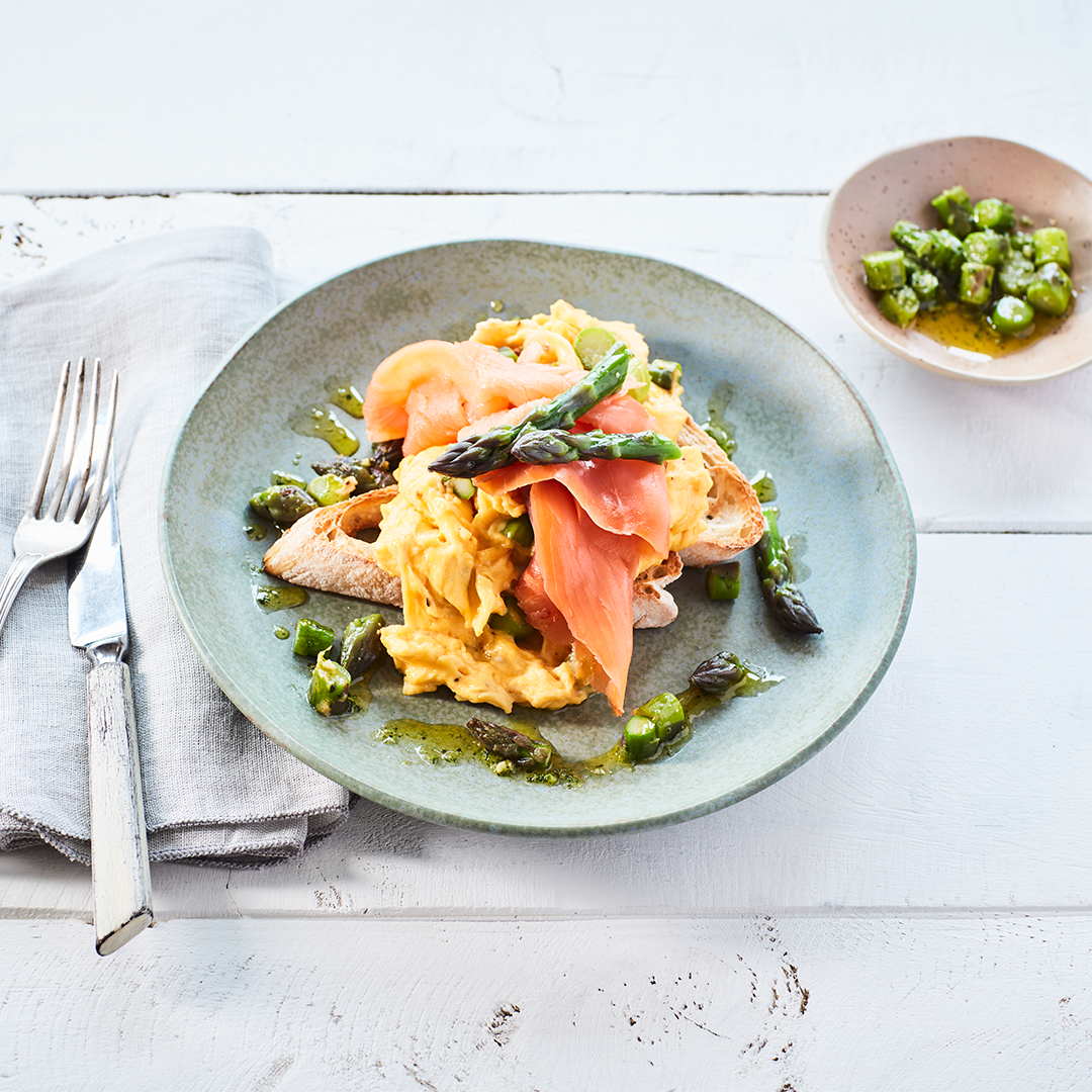 Scrambled eggs on toast with salmon and asparagus, contained in a bowl next to cutlery napkin