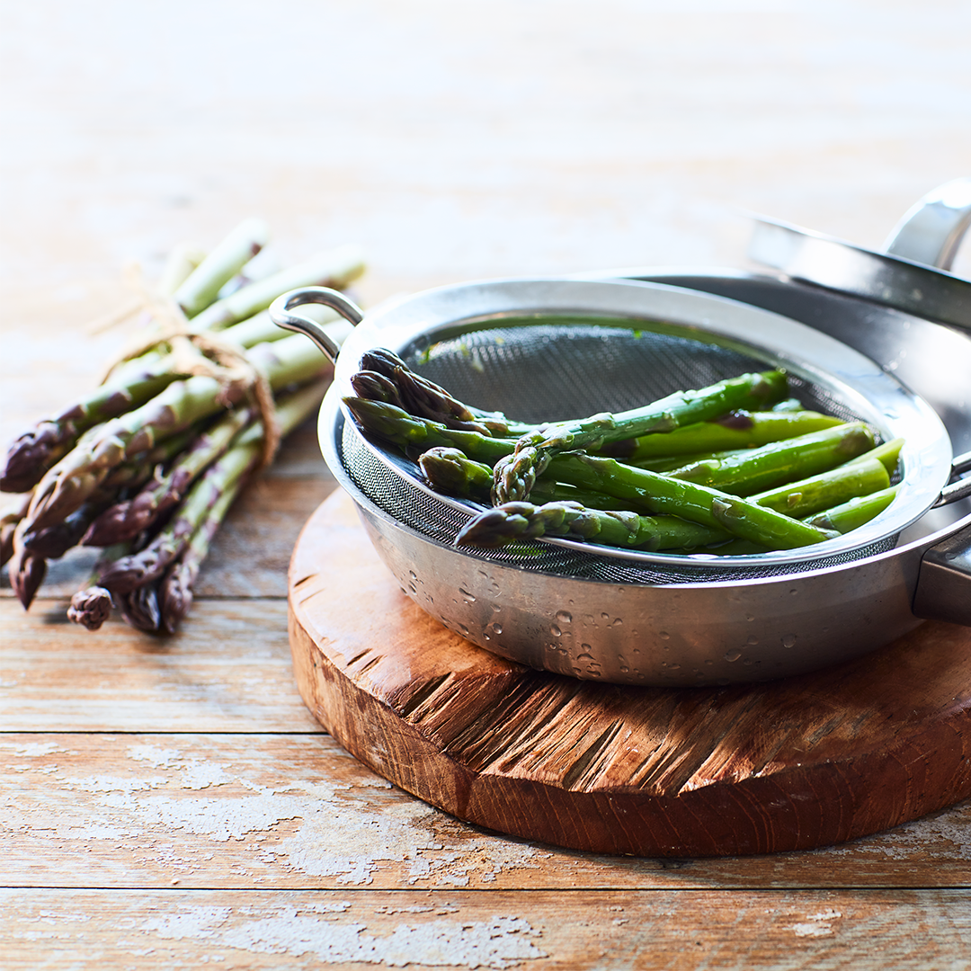 Multiple steamed asparagus sitting in a sieve on a circular wooden block